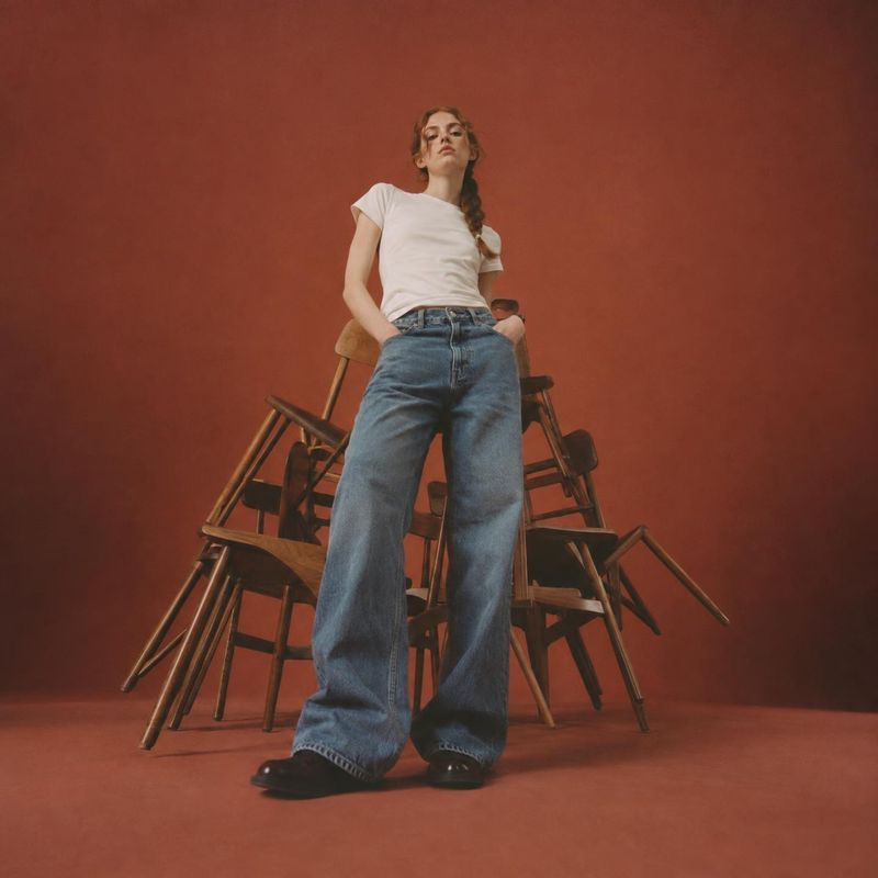 wide-leg jeans look book product photo: A model poses in wide-leg blue jeans and a white T-shirt among stacked wooden chairs against a warm terracotta studio b…