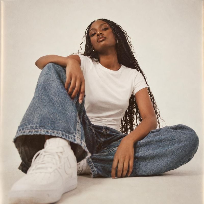 sneakers look book product photo: A model in a white tee, relaxed blue jeans and white Nike sneakers sits casually against a warm, pale studio backdrop, with s…