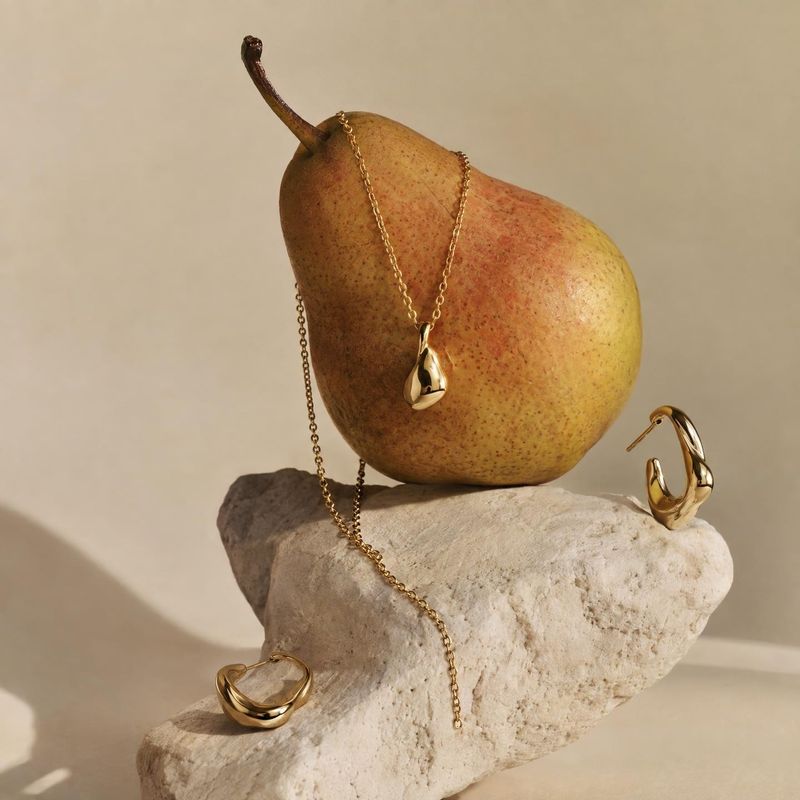 gold jewelry set still life product photo: Gold-toned necklace and sculptural earrings are artfully arranged with a ripe pear on a pale stone against a warm ne…