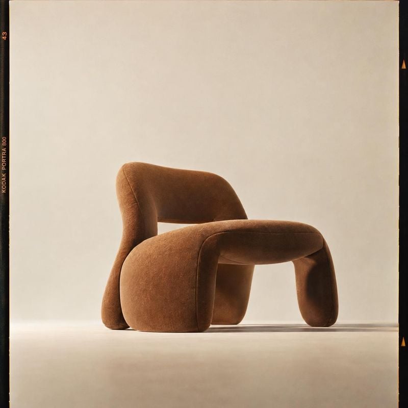 upholstered lounge chair packshot product photo: A sculptural brown upholstered lounge chair sits alone against a warm cream studio backdrop, softly lit with g…