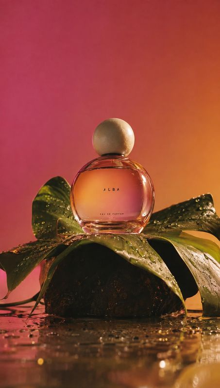 perfume still life product photo: A rounded glass perfume bottle rests among glossy, dew-covered leaves on a wet surface, lit by warm pink and amber gradients…