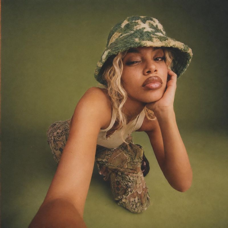 camouflage bucket hat and coordinated outfit look book product photo: A model in a textured camouflage bucket hat and coordinated outfit leans toward the camer…
