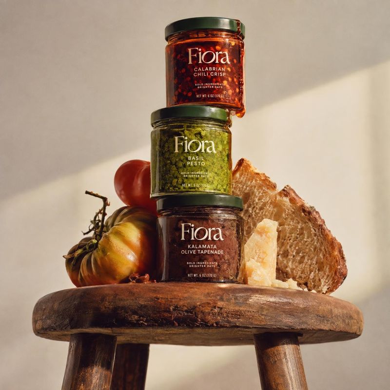 three jars of spreads and condiments ingredients product photo: Three labeled jars of chili crisp, basil pesto, and olive tapenade are stacked on a rustic wood…