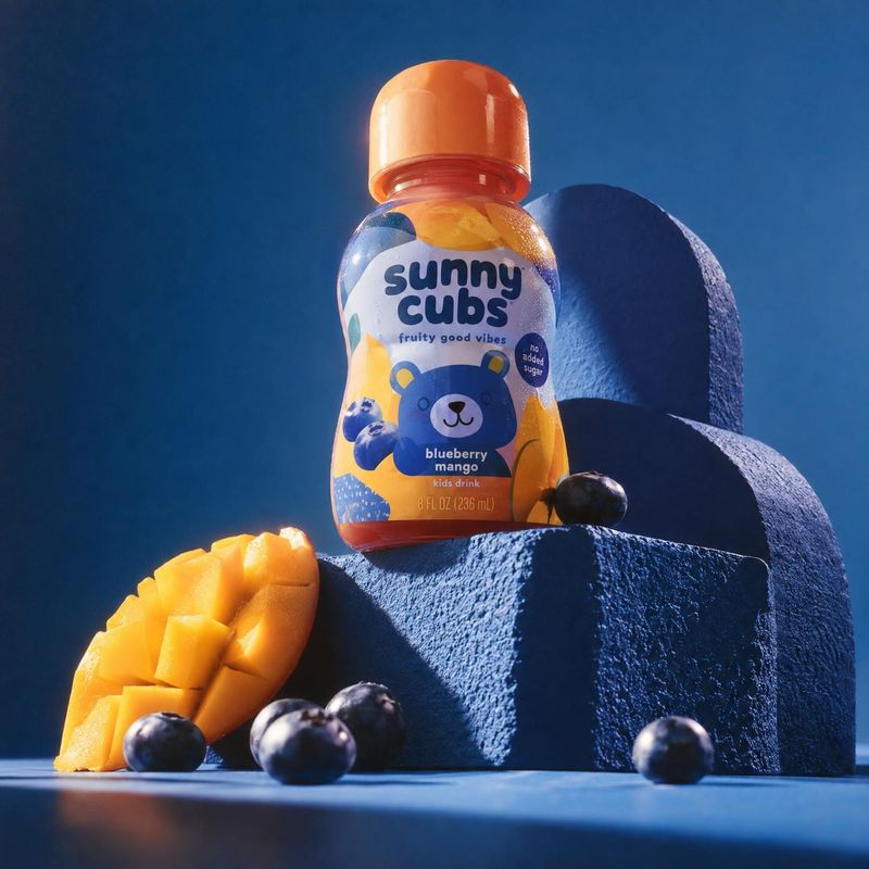 kids fruit drink bottle ingredients product photo: A colorful kids’ fruit drink bottle sits among mango pieces and blueberries on sculptural blue forms, lit wi…