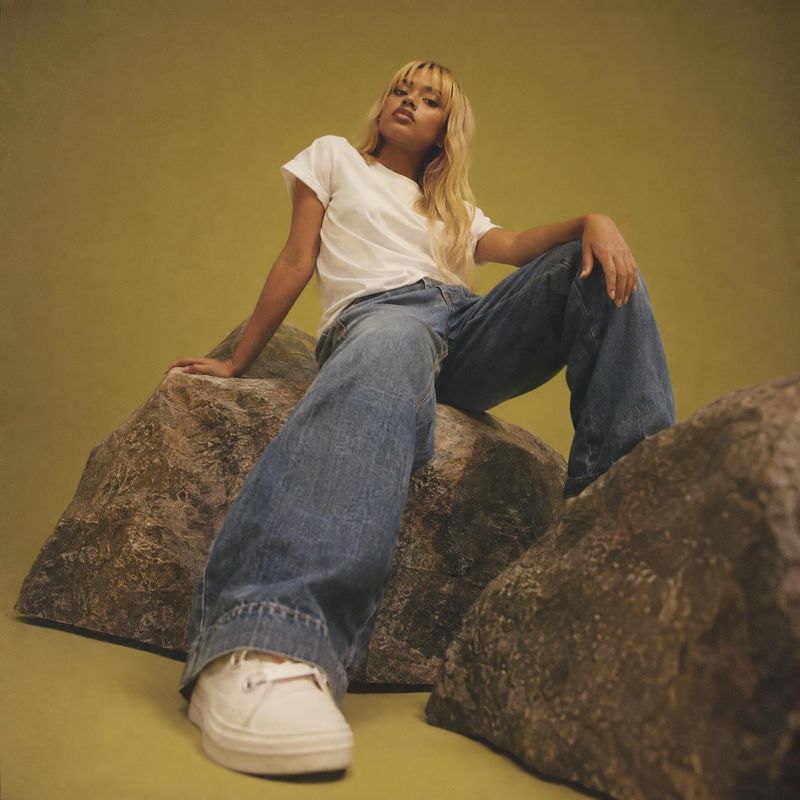 wide-leg jeans look book product photo: A model in a white tee, loose wide-leg jeans, and pale sneakers poses on sculptural rocks against a muted olive studio…