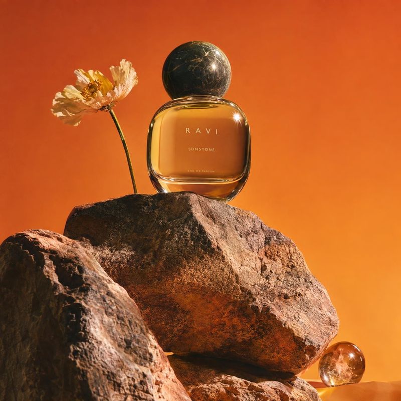 perfume still life product photo: A glass perfume bottle labeled RAVI SUNSTONE stands among rugged rocks beside a pale flower, lit warmly against a vivid orang…