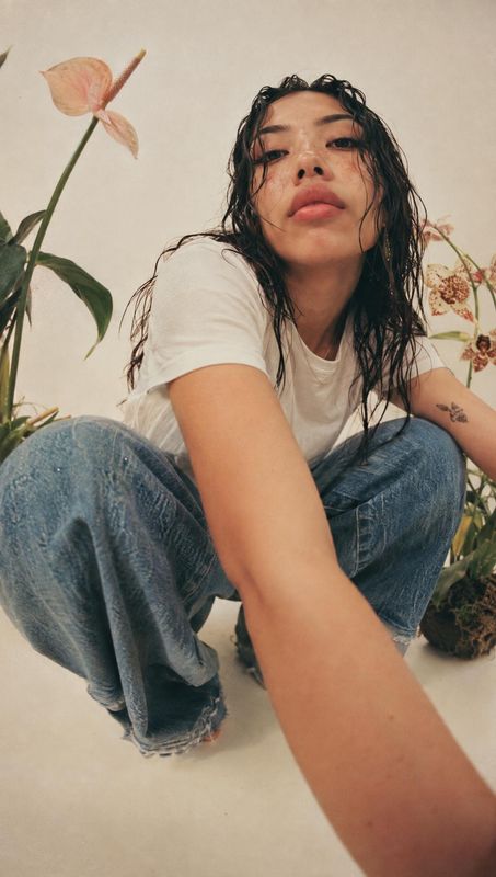 casual outfit look book product photo: A model in a white T-shirt and relaxed blue jeans crouches among potted flowers against a warm neutral studio backdrop,…