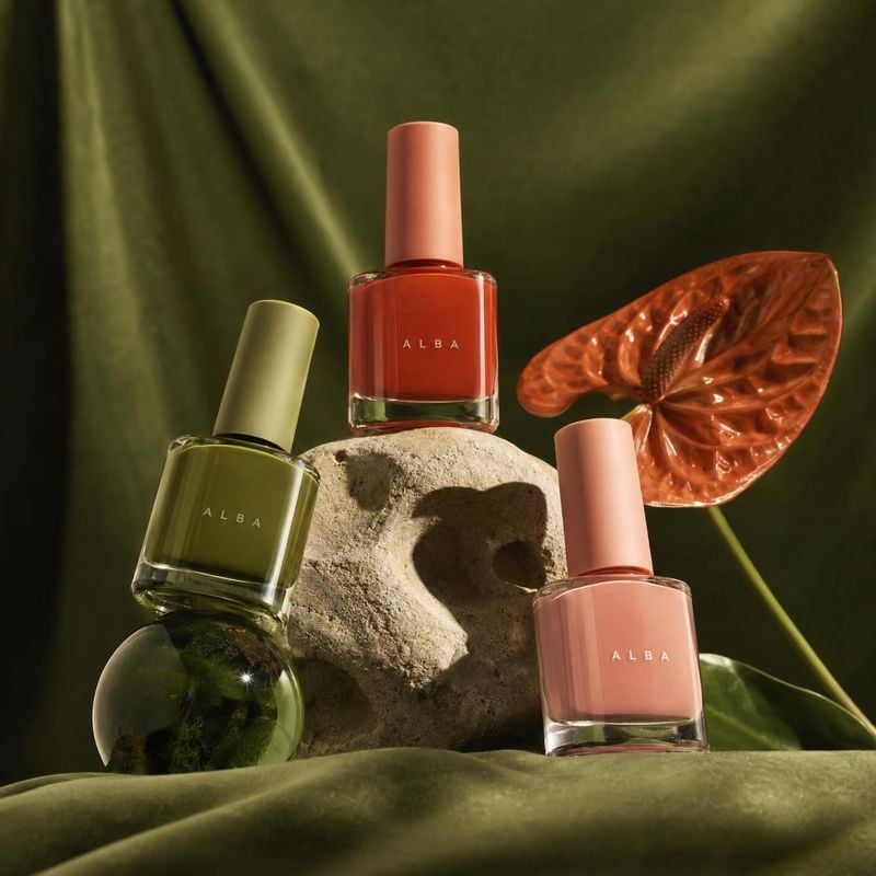 nail polish still life product photo: Three glass nail polish bottles in olive, coral, and blush tones are arranged on a sculptural stone among glossy botanica…