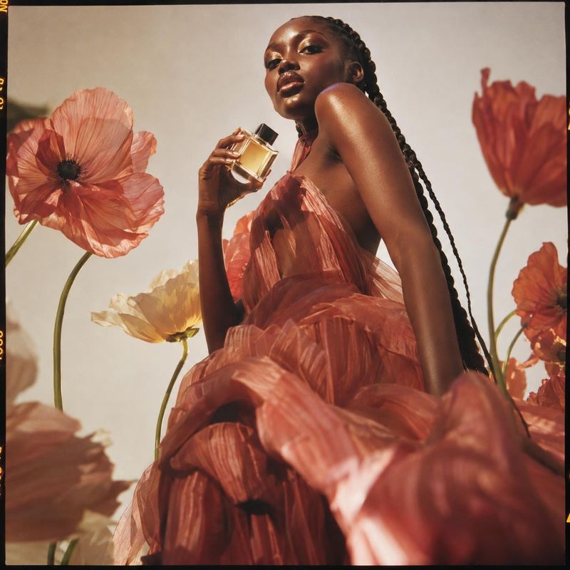 perfume look book product photo: A model in a voluminous coral gown holds a perfume bottle among oversized poppies, captured in warm sunlight with a dramatic,…