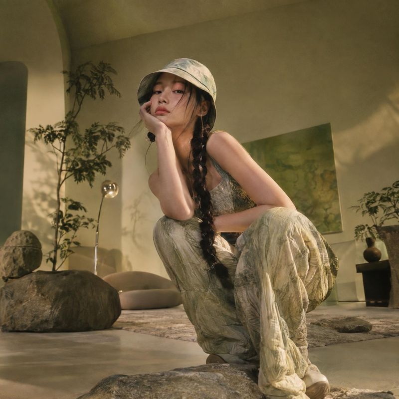 fashion outfit look book product photo: A woman in a softly patterned outfit and bucket hat poses low to the ground in a warm, plant-filled interior, lit by ge…