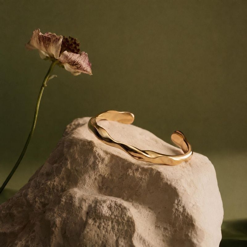gold cuff bracelet close-up detail product photo: A sculptural gold cuff rests on a pale textured stone beside a dried flower, softly lit against an olive-gree…