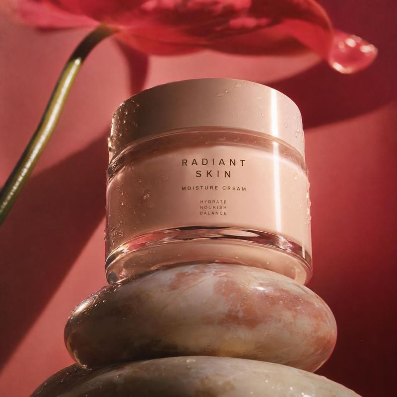 moisture cream still life product photo: A moisture cream jar rests on smooth stacked stones against a saturated red backdrop, with a flower above it and warm…