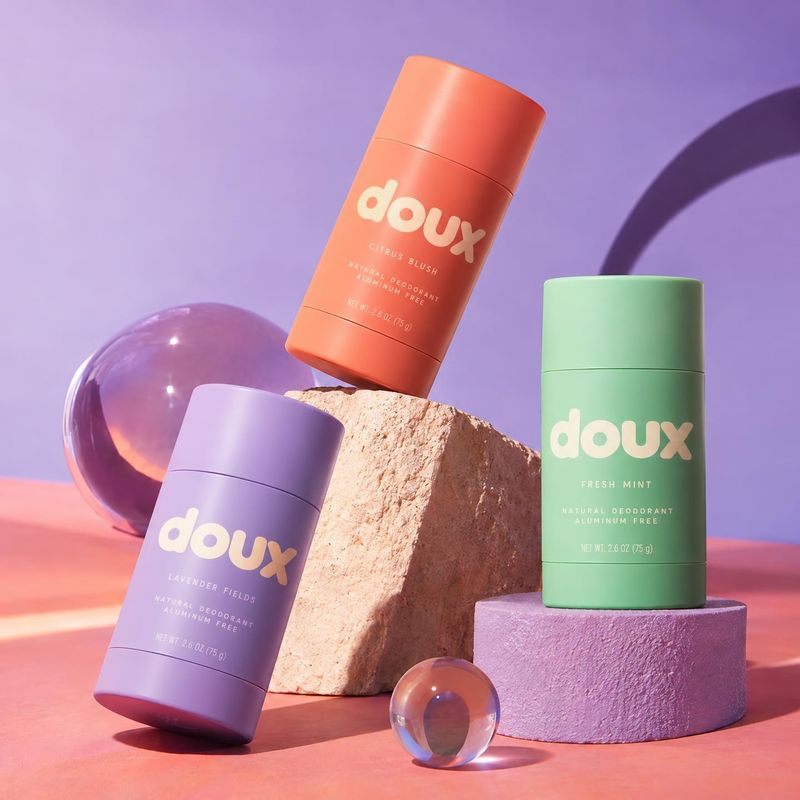 deodorant sticks group lineup product photo: Three colorful deodorant sticks are arranged on sculptural blocks with glass spheres against a pastel lavender and…