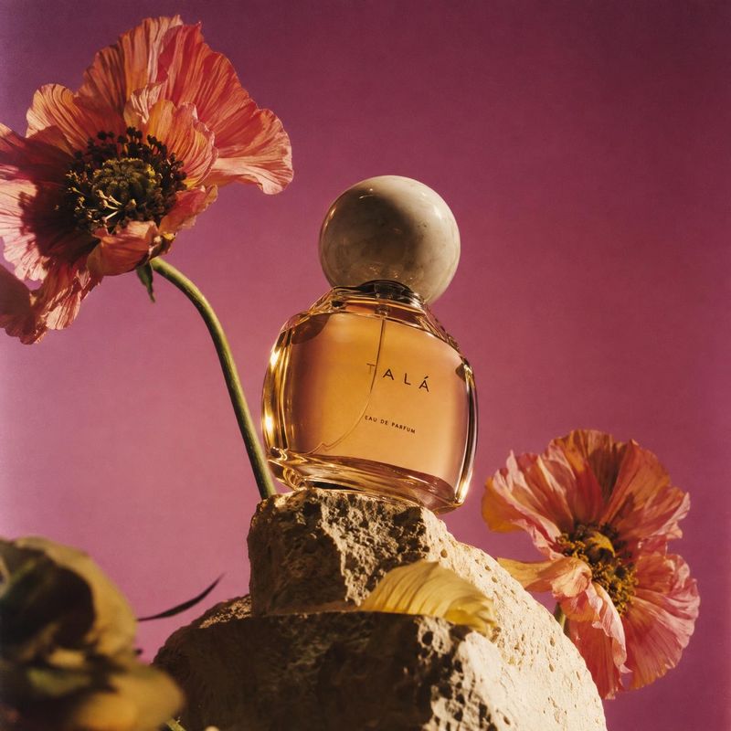perfume still life product photo: An amber perfume bottle with a rounded cap sits on a rugged stone among orange flowers against a rich pink backdrop, lit with…