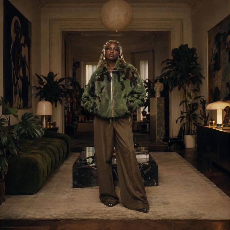 camouflage fleece jacket look book product photo: A model wears an oversized green camouflage fleece jacket and brown trousers in a warmly lit, plant-filled li…