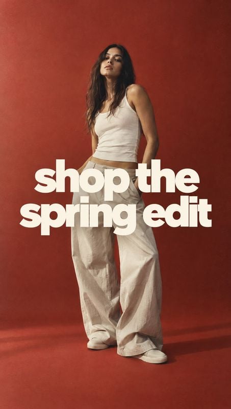 women's spring outfit model try-on product photo: A model poses in a white tank top and loose trousers against a warm red studio backdrop, with bold cream lett…