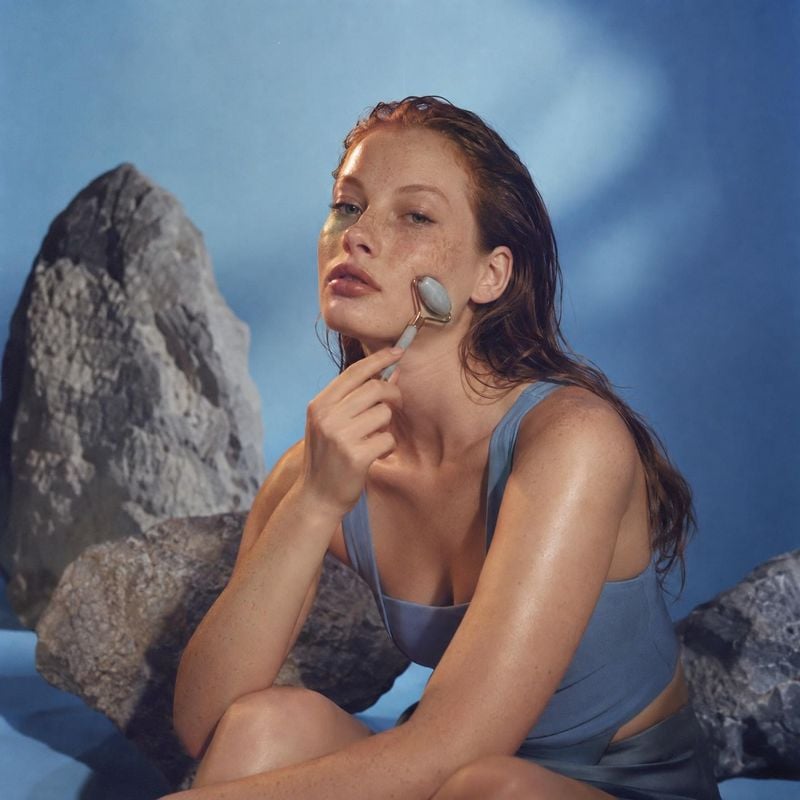 facial roller model try-on product photo: A woman in blue activewear gently rolls a facial massage tool along her cheek against a sculptural rocky set and cool…