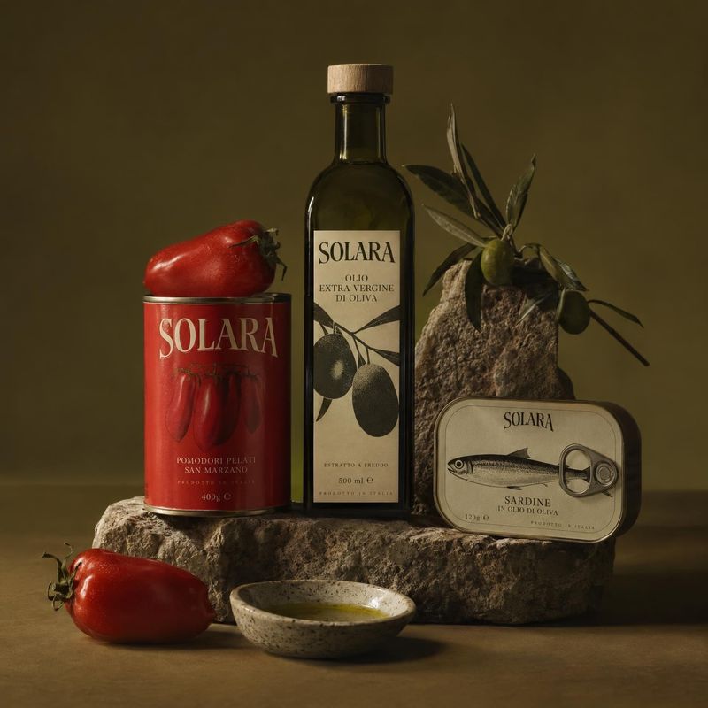 Italian food products still life product photo: A bottle of olive oil, canned tomatoes and sardines are arranged with fresh tomatoes, olive branches and a smal…