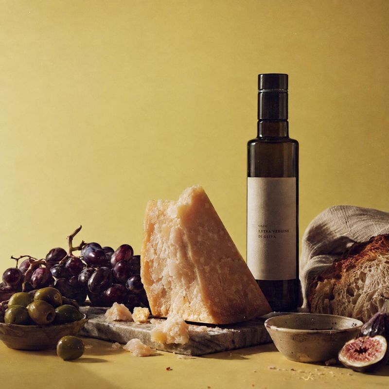 olive oil ingredients product photo: A dark glass olive oil bottle is styled with olives, cheese, bread, and figs against a warm yellow backdrop, creating a ri…