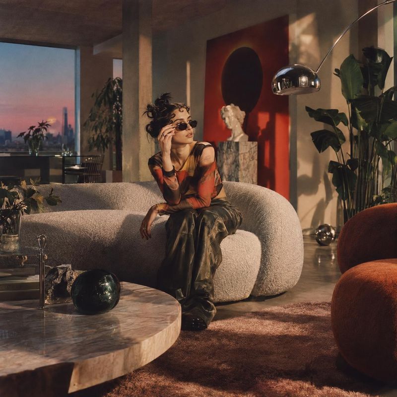 sunglasses look book product photo: A woman in narrow sunglasses and a sheer patterned top lounges on a sculptural sofa in a warmly lit modern interior, with s…