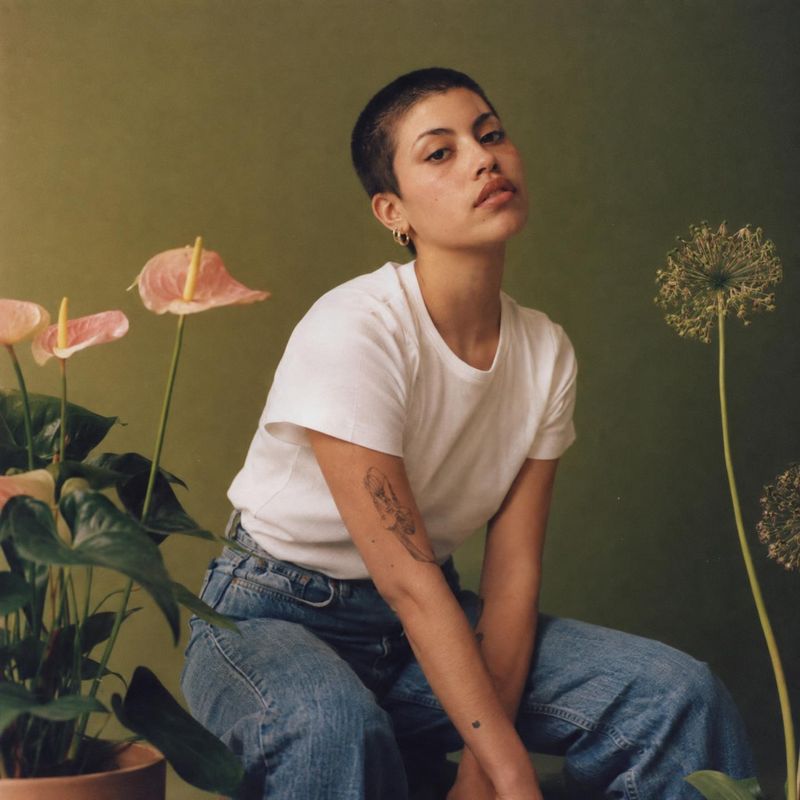 white T-shirt and jeans look book product photo: A seated model in a white T-shirt and blue jeans poses among pink flowers against an olive-green studio backdr…