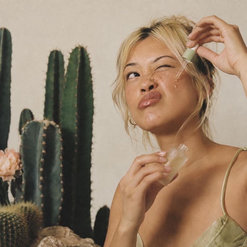 facial serum beauty close-up product photo: A close beauty portrait shows a blonde woman applying facial serum with a dropper beside tall cacti, in warm natura…
