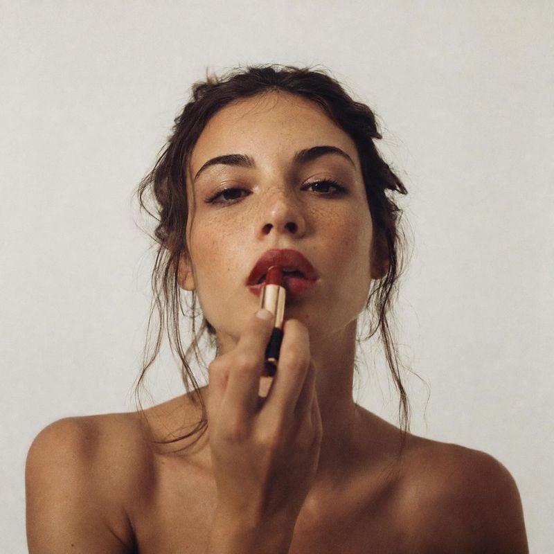 lipstick beauty close-up product photo: A woman applies deep red lipstick in a close studio portrait against a softly lit neutral backdrop, creating an intimat…