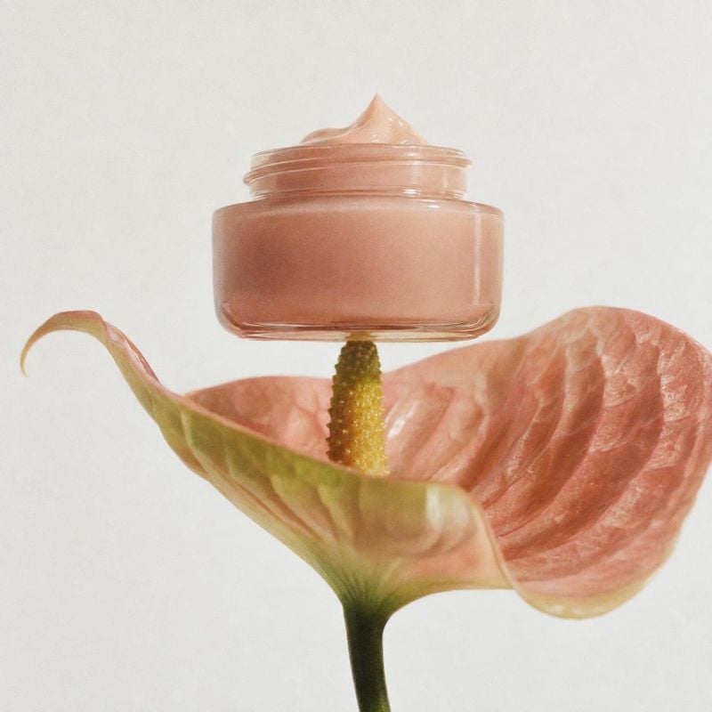 face cream beauty product photo: A peach-toned cream jar rests atop a pink anthurium against a pale seamless backdrop, styled with soft light for a delicate bo…