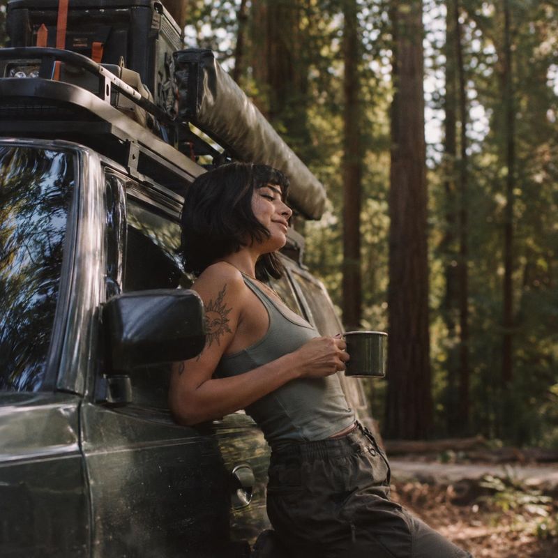 off-road vehicle vehicle exterior product photo: A woman in casual outdoor clothing leans against a gear-laden off-road vehicle in a forest, holding a metal mu…