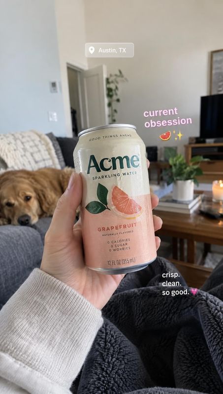 grapefruit sparkling water can lifestyle scene product photo: A hand presents a grapefruit sparkling water can in a cozy living room, with a dog on the sofa an…