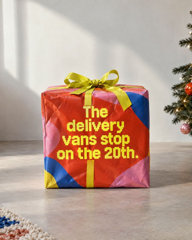 gift-wrapped package lifestyle scene product photo: A brightly wrapped gift with a lime-green bow sits on a sunlit floor beside a decorated Christmas tree, wit…