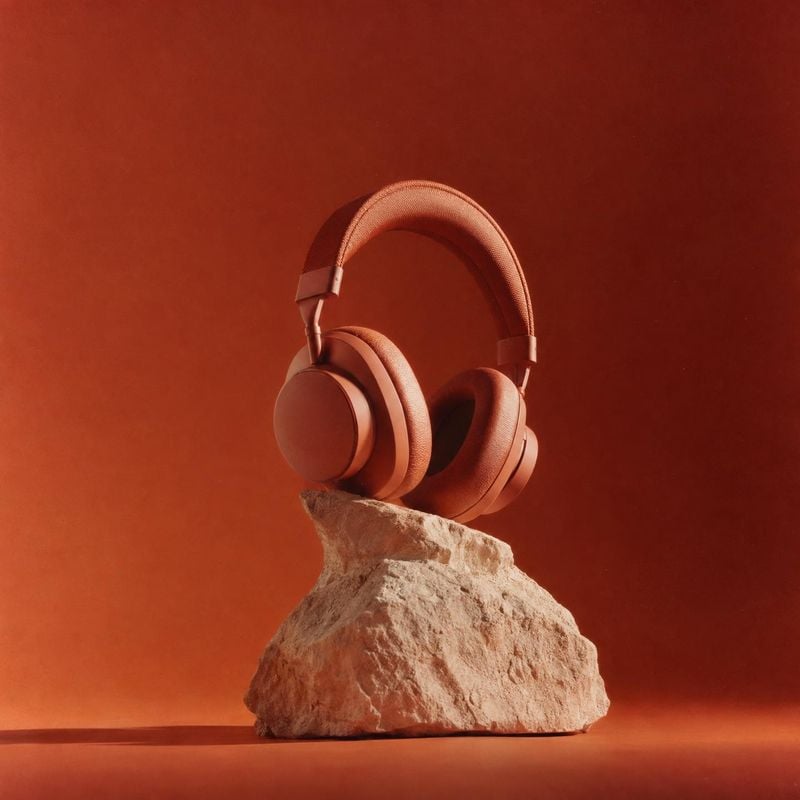 over-ear headphones electronics product photo: A pair of warm terracotta-colored over-ear headphones rests on a rough stone against a seamless burnt-orange stu…
