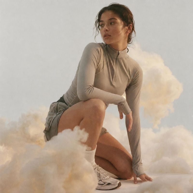 athletic outfit look book product photo: A woman in a muted athletic outfit crouches among soft clouds on a pale studio set, lit with warm, diffused light for…