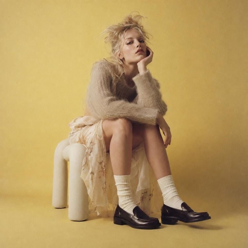 black loafers look book product photo: A blonde model poses seated on a sculptural stool against a warm yellow studio backdrop, wearing a soft knit sweater, fl…