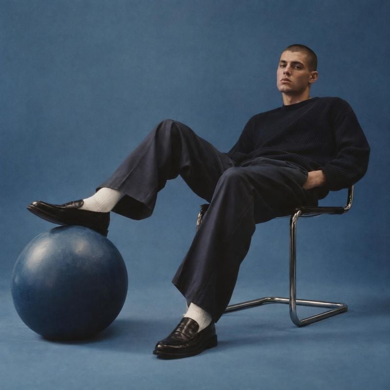 men’s clothing and loafers look book product photo: A male model in a dark knit top, tailored trousers, white socks and loafers reclines on a chrome chair agai…
