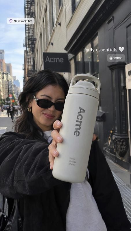 insulated water bottle lifestyle scene product photo: A woman in sunglasses presents a large white insulated bottle on a New York sidewalk, with the storefront…