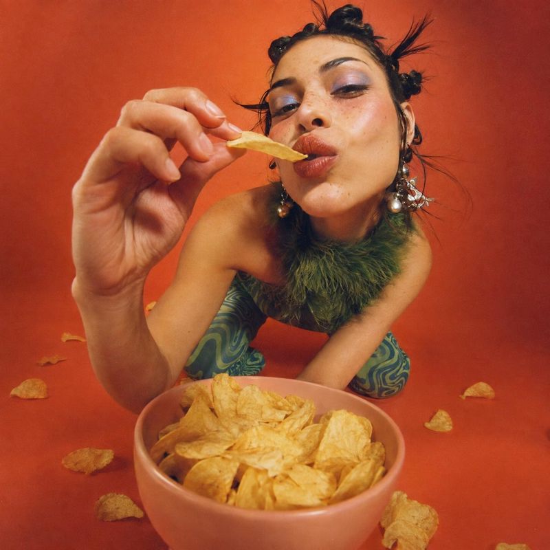 potato chips look book product photo: A fashion-styled woman leans toward the camera to eat a potato chip above a bowl of chips, set against a vivid orange stu…
