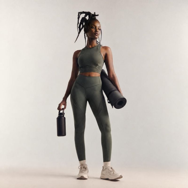 activewear set model try-on product photo: A model poses in a dark green athletic top and leggings, holding a yoga mat and water bottle against a softly lit ne…