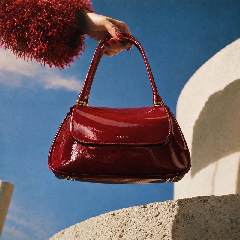 burgundy handbag product in hand product photo: A glossy burgundy handbag is presented by a manicured hand against a vivid blue sky and sunlit stone, creating…