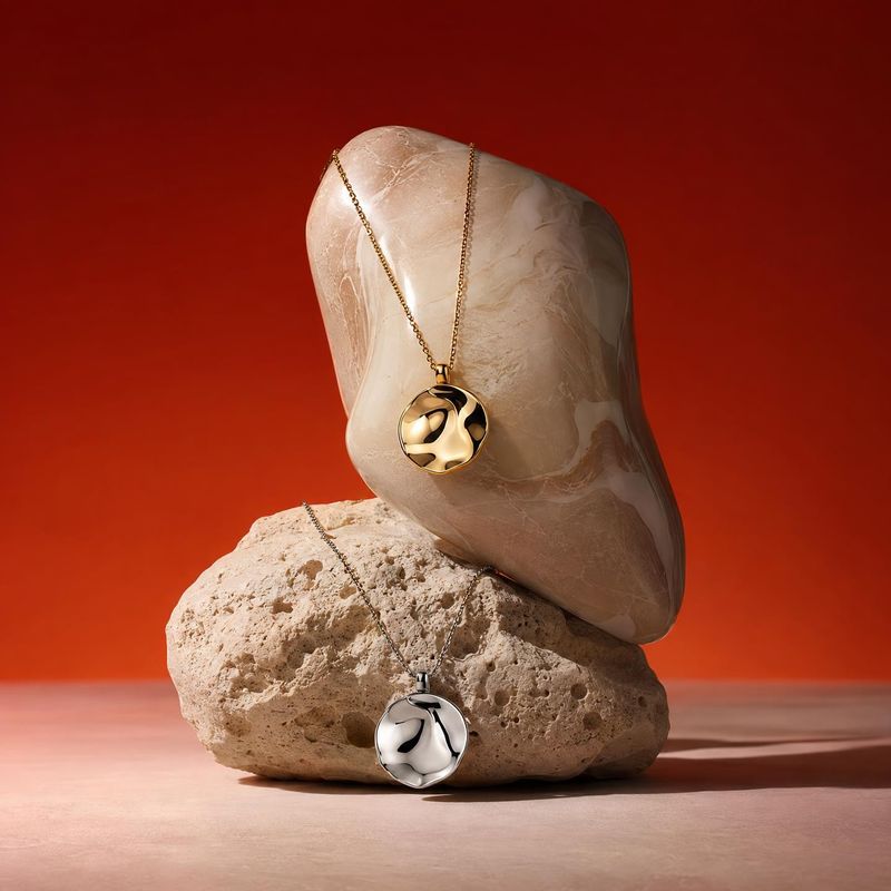 pendant necklaces jewelry product photo: Two sculptural gold- and silver-tone pendant necklaces are draped over pale natural stones against a vivid red studio…