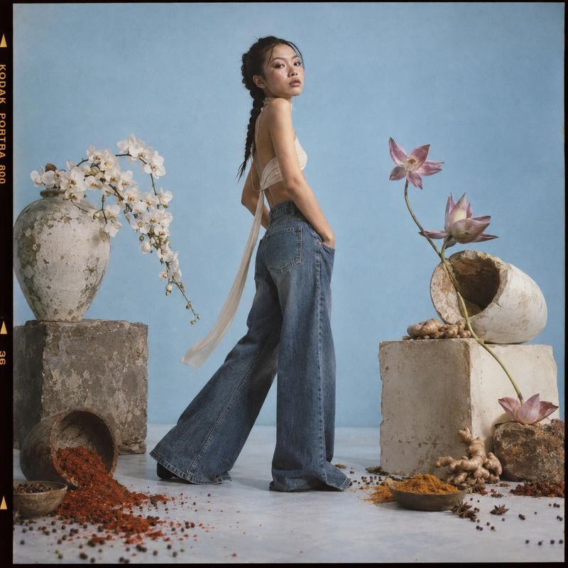 wide-leg jeans look book product photo: A model poses in a white top and wide-leg jeans against a pale blue studio backdrop, framed by sculptural stone vessels…