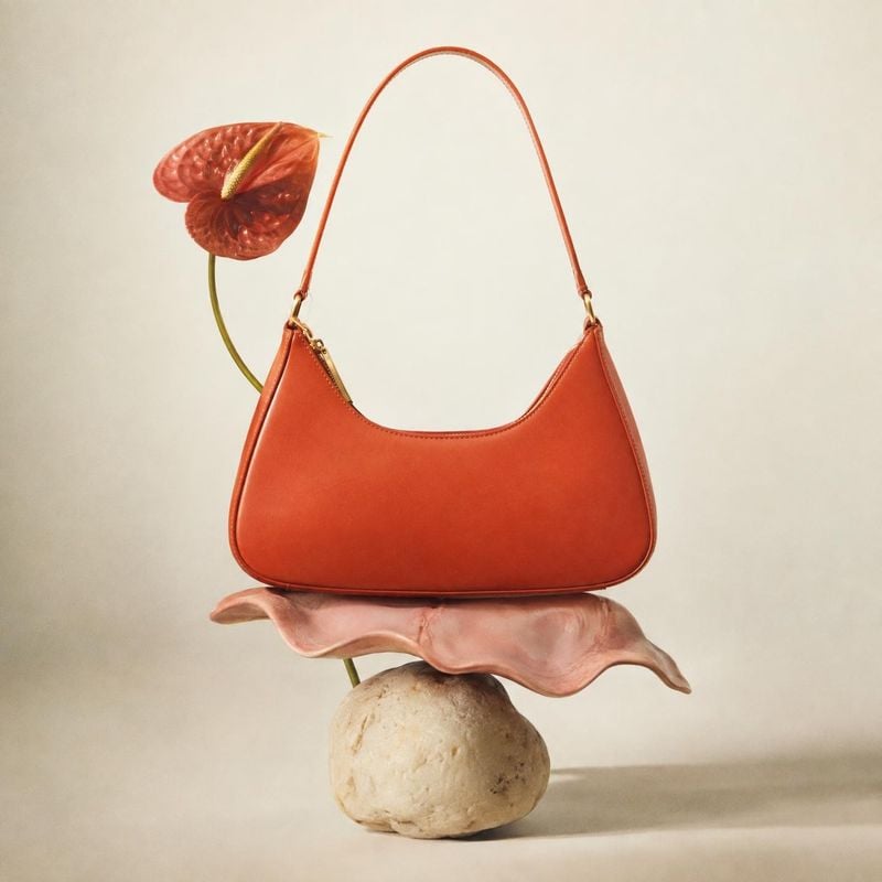 handbag jewelry product photo: A sculptural orange-red handbag is balanced on a pink shell and pale stone beside a red anthurium, photographed against a softly…