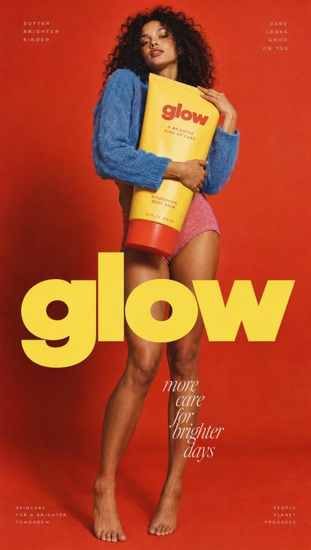 brightening body balm look book product photo: A curly-haired woman in a blue sweater and pink shorts holds a yellow body balm against a vivid red studio backd…
