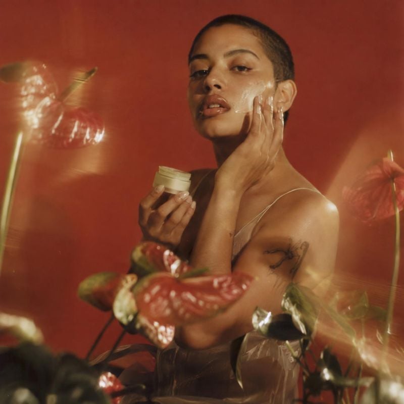 face cream model try-on product photo: A model holds a small jar of face cream and applies it to her cheek against a warm red studio backdrop, with glossy bota…