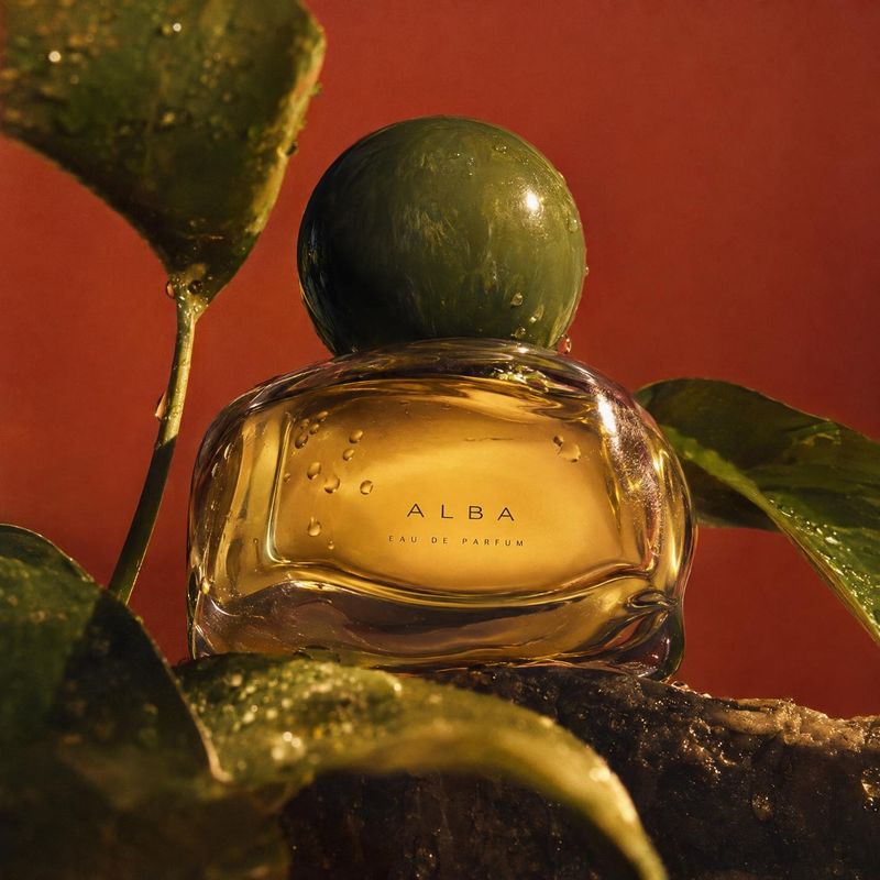 perfume close-up detail product photo: A golden perfume bottle with a dark green rounded cap sits among glossy leaves against a warm red backdrop, lit with dra…