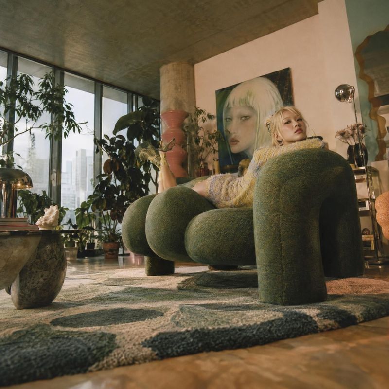 sculptural upholstered armchair room set product photo: A blonde woman reclines in a rounded moss-green armchair in a leafy modern interior, where warm natural…