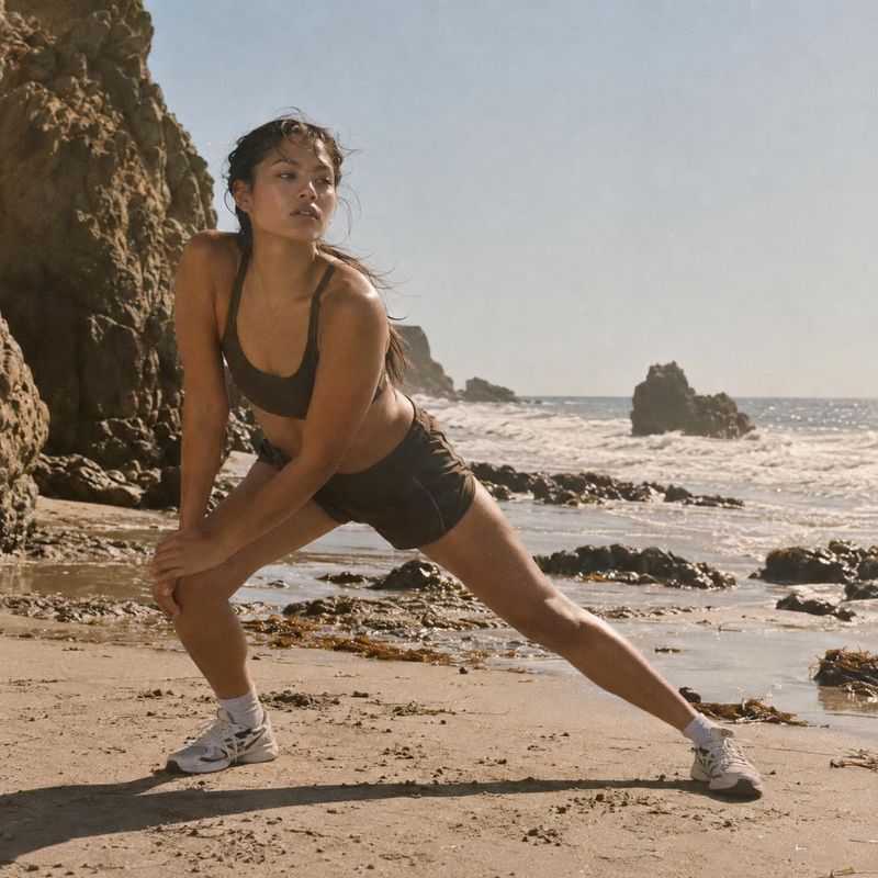 athletic wear model try-on product photo: A woman in dark athletic wear stretches on a rocky beach, with warm natural sunlight, open sky and a relaxed active m…