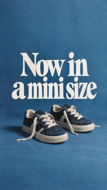 children's sneakers still life product photo: A pair of small navy canvas sneakers with white laces sits on a blue studio surface beneath large cream display l…