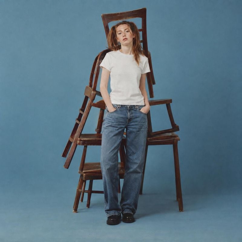 jeans and T-shirt look book product photo: A model in a white T-shirt, loose blue jeans and dark shoes stands in front of stacked wooden chairs against a blue…