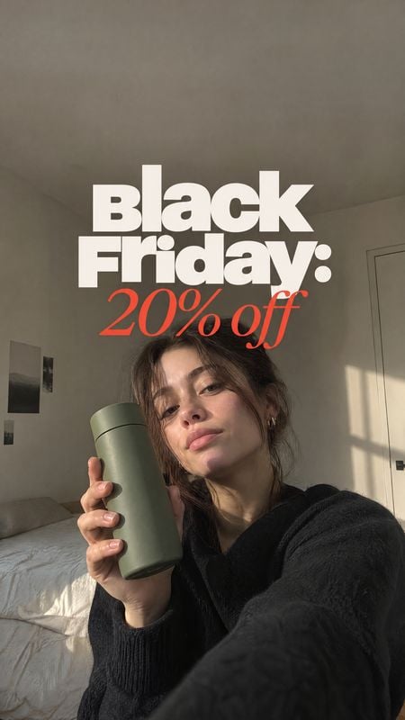 reusable tumbler sports product photo: A woman in a black sweater holds a muted green tumbler for a bedroom selfie, with warm sunlight and bold Black Friday di…
